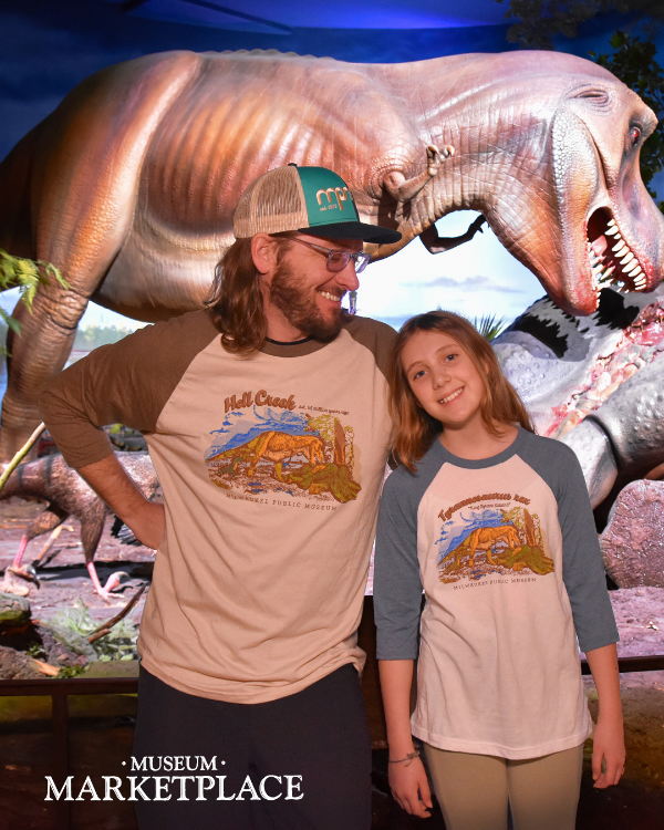 A father and daughter wearing Hell Creek T. rex t-shirts posing in front of the Hell Creek exhibit